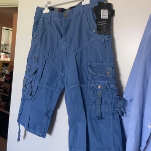 Blå cargoshorts från Adowops - Snygga blå cargoshorts med flera stora fickor och coola detaljer som knappar och snörning vid benslut. Tillverkade i 100% bomull för en skön känsla. Perfekta för dig som gillar en avslappnad och funktionell stil.Helt nya med etikett 