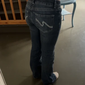 Mörkblå bootcut jeans med broderi - Säljer ett par mörkblå jeans med bootcut-modell och snygga broderade detaljer på bakfickorna. Jeansen har låg midja och klassisk femficksdesign. Perfekta för dig som gillar en tidlös och avslappnad look.