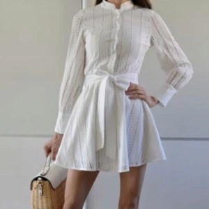 Vit broderad kortklänning med knyt - Superfin vit kortklänning i bomull med broderade detaljer och knytband i midjan. Klänningen har långa ärmar, ståkrage och knappar framtill. Perfekt för sommarens alla tillfällen.