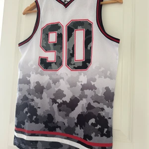 Basketlinne Tenth camo svart/vit - Snyggt basketlinne från Tenth i vitt med svart och grått camouflagemönster och stora siffror 90 på framsidan. Röd och svart rand runt hals och ärmhål. Tillverkat i luftig polyester, perfekt för träning eller sport. Storlek 164-170. 