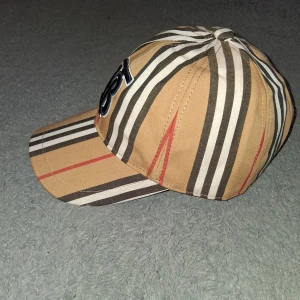 Randig keps från Burberry - Snygg keps från Burberry med klassiskt randigt mönster i beige, svart, vitt och rött. Framtill finns en broderad svart och vit logga. Kepsen har justerbar knäppning bak och är tillverkad i bomull med detaljer i kalvskinn. Perfekt för dig som vill sticka ut med stil.