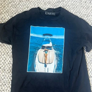 Svart t-shirt med båttryck från L'ARTE - Svart t-shirt från L'ARTE med ett stort färgglatt tryck på bröstet som visar en person som solar på en båt mitt ute på havet. Klassisk rund halsringning och korta ärmar. Perfekt för dig som gillar statement-plagg och coola motiv.