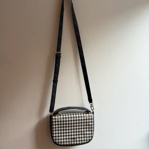 Svartvit axelväska från Guess - Snygg axelväska från Guess med klassiskt svartvitt hundtandsmönster. Väskan har justerbar axelrem, dragkedja och flera fack inuti. Materialet är tålig syntet med Guess-logga i metall framtill. Perfekt storlek för det viktigaste och en riktigt trendig detalj.