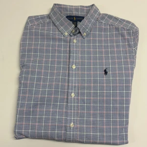 Rutig skjorta från Ralph Lauren XL - Klassisk rutig skjorta från Ralph Lauren i blått, vitt och röda detaljer. Skjortan har lång ärm, knappar framtill och broderad logga på bröstet. Tillverkad i mjuk bomull och passar perfekt till en avslappnad men ändå stilren look. Storlek XL 