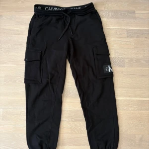 Svarta mjuka cargopants från Calvin Klein - Svarta cargopants från Calvin Klein med elastisk midja och snörning. Byxorna har stora fickor på sidorna och CK-logga på ena benet. Tillverkade i mjukt material för chill och avslappnad känsla. 