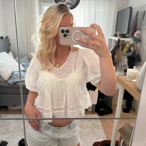 Vit blus med puffärmar och spetsdetaljer - Supersöt vit blus med korta puffärmar och fina spetsdetaljer. Den har en lös och luftig passform, rund halsringning och volangliknande nederkant. Perfekt för varma dagar och ger en romantisk vibe till outfiten.