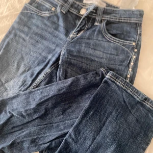Blå jeans med nitar från Bershka - Snygga blå jeans från Bershka med coola detaljer som nitar och beige broderi på bakfickorna. Klassisk femficksmodell med diamant knappar och kontrastsömmar. Perfekta för dig som vill sticka ut lite extra.