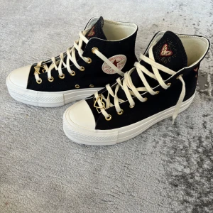 Converse svart high-top med hjärtdetalj - Svarta Converse high-top sneakers med vit platt sula och rund tå. Skorna har vita snören, guldfärgade öljetter och en unik hjärtformad detalj på snörningen. Tungan pryds av ett broderat rött hjärta med gulddetaljer. Klassisk Converse-logga på sidan. Materialet är bomull.