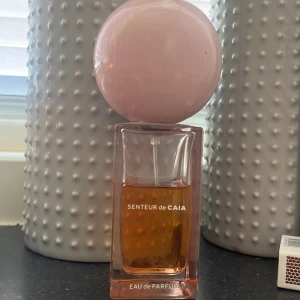 Senteur de Caia Eau de Parfum - Snygg parfymflaska från Caia med en unik design – rektangulär rosa glasflaska och en stor rund ljusrosa kork. Innehållet är en amberfärgad Eau de Parfum som känns lyxig och modern. Perfekt för dig som gillar trendiga och stilrena detaljer på sminkbordet.