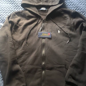 Svart hoodie från Polo Ralph Lauren - Svart hoodie från Polo Ralph Lauren med dragkedja och huva. Klassisk broderad logga på bröstet och snörning i huvan. Mjuk bomullskänsla och känguruficka framtill. Perfekt för en chill och stilren look.
