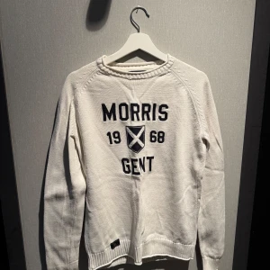 Vit stickad tröja Morris Gents S - Vit stickad tröja från Morris Gents med svart broderad text och emblem framtill. Klassisk rund halsringning och långa ärmar. 