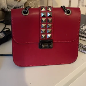 Röd axelväska med nitar och kedja - Cool röd axelväska i skinnimitation med silverfärgade nitar framtill och en snygg kedjerem. Väskan har en fyrkantig form och stängs med ett metallspänne. Perfekt för dig som vill sticka ut med en edgy accessoar.