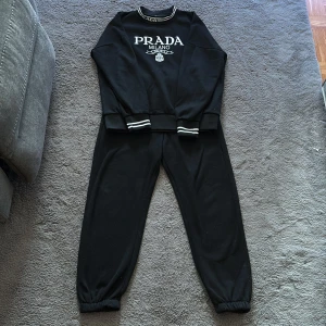 Svart sweatshirt från Prada Milano - Svart sweatshirt från Prada Milano med vit logga framtill och coola ränder på muddar och krage. Tröjan har lång ärm och relaxed fit, perfekt för en chill look. Materialet är mjukt och känns skönt mot huden. Snygg detalj med broderad logga och ribbade muddar.