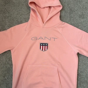 jättesöt rosa hoodie från gant - Snygg ljusrosa hoodie från GANT med broderad logga och sköld. Tröjan har huva, magficka och långa ärmar. Perfekt för dig som gillar en avslappnad och trendig stil. Tillverkad i mjuk bomull för extra komfort.