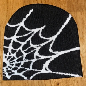 Svart mössa med vitt spindelnätsmönster - Cool svart mössa med ett stort vitt spindelnätsmönster som täcker hela ytan. Perfekt för dig som vill sticka ut med en edgy och unik stil. Mössan är stickad och sitter tajt på huvudet. Passar både till höst och vinter.