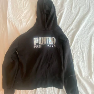 Svart hoodie från Puma - Svart hoodie från Puma med stort holografiskt logotryck på bröstet och coola symboler under. Tröjan har huva och långa ärmar, perfekt för en chill och sportig look. Mjuk insida och klassisk passform. Pris kan diskuteras.