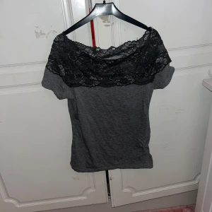 Grå topp med svart spetsdetalj - Snygg grå topp med bred svart spets upptill som går över axlarna. Offshoulder-modell med korta ärmar och mjukt material. Perfekt för dig som gillar en stilren look med lite extra detalj.står ingen storlek men skulle gissa på en S.