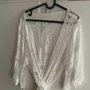 Vit lång spetskappa  - Superfin lång vit spetskapoa i transparent spets med broderade detaljer och omlottmodell. Klänningen har vida trekvartsärmar och passar med ett valfritt bälte / knyt i midjan för en snygg siluett eller kaxigt bara öppen. Perfekt för sommaren eller inomhus till alla tillfällen! Funkar med både kjol och leggings eller bara bikini. 