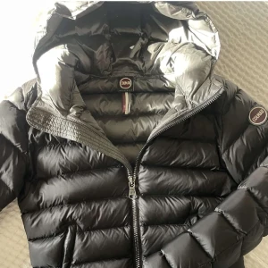 Svart dunjacka från Colmar - Snygg svart dunjacka från Colmar med dragkedja och huva. Jackan har en quiltad design och en rund logotyp på ärmen. Perfekt för kyligare dagar. Jackan har ett litet hål i innerfickan. Men inget som varken syns när den är på eller stör andvändningen. Passar xs/s bilderna är inte mina men det är den jackan bilderna där ja har på mig jackan är min egna bild d  ett är även småt hål på ryggen alla bilder är lånade förutom den andra och sista bilden