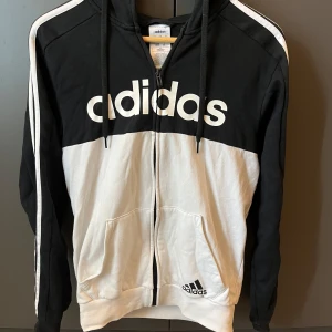Svartvit Adidas hoodie med dragkedja XS - Svart och vit hoodie från Adidas i storlek XS. Klassisk design med stor logga framtill, vita ränder längs ärmarna och huva med snörning. Dragkedja hela vägen och två fickor framtill. Perfekt för en sportig och avslappnad stil.