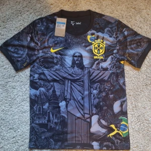 Brasil "Christ the redeemer" Tröja - Unik svart Brasilien fotbollströja från Nike med coolt grafiskt tryck av Kristusstatyn och brasilianska symboler. Gula detaljer med CBF-emblem och Nike-logga. Tillverkad i Dri-FIT-material som andas. Perfekt för dig som vill sticka ut på planen eller läktaren.