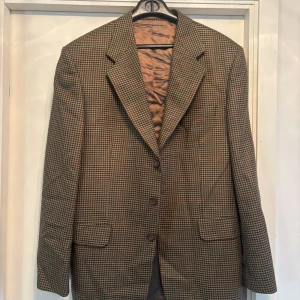 Rutig kavaj från Oscar Jacobson - Snygg rutig kavaj från Oscar Jacobson i klassisk brun och beige ton. Kavajen har tre knappar framtill, två fickor med lock och en stilren krage. Fodret är brunt och känns lyxigt. Perfekt för dig som vill ha en tidlös och cool look.