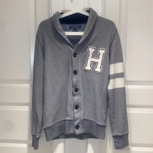 Grå knapp-kofta från Tommy Hilfiger - Snygg grå kofta från Tommy Hilfiger i vintage fit med stora vita H-trycket på bröstet och två vita ränder på ena ärmen. Koftan har knappar hela vägen och en skön krage. Perfekt för dig som gillar college-vibe och klassisk stil.