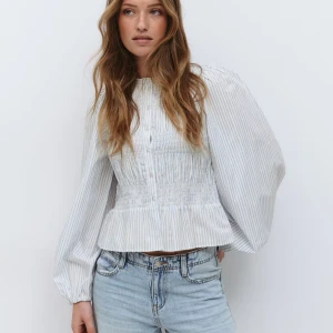Randig blus med puffärm från Gina Tricot - Supersöt blus i vitt med tunna blå ränder, puffiga långärmar och smockad midja. Rund halsringning, knappar framtill och volang nedtill🩵har små sminkfläck vid kragen