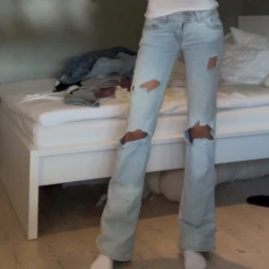 Ljusblå lågmidjade jeans med slitningar - Säljer ett par lowwaist ljusblå bootcut jeans med snygga slitningar på knäna och låren. Jeansen har låg midja och klassisk femficksmodell. Perfekta för dig som gillar en avslappnad och trendig look.
