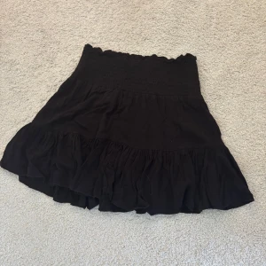 Svart volangkjol med smock - Supersöt svart kortkjol med bred smock i midjan och volang nertill. Kjolen har en luftig känsla och passar perfekt till sneakers. Enkel att styla.