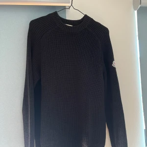 Svart stickad tröja från Moncler - Säljer en svart stickad tröja från Moncler med rund halsringning och ribbade muddar. Tröjan har lång ärm och Moncler-logga på ena ärmen. Perfekt för dig som gillar stilren och klassisk design.