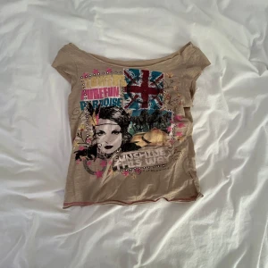 Beige topp med grafiskt tryck - Cool beige topp med korta ärmar och ett stort grafiskt tryck framtill med färgglada detaljer, text och motiv av ett ansikte och brittiska flaggan. T-shirten har en avslappnad passform och är tillverkad i mjukt bomullsmaterial. Perfekt för dig som gillar unika statement-plagg.