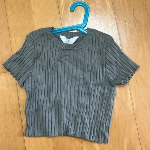 Grå ribbad topp från H&M - Kortärmad grå topp från H&M med ribbad struktur och rund hals. Toppen har en croppad passform och vågiga kanter vid ärmslut och nederkant. Perfekt att styla med jeans eller kjol för en chill look.