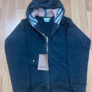 Svart hoodie från Burberry med dragkedja - Svart hoodie från Burberry i storlek M med klassisk dragkedja framtill och huva med ikoniskt rutigt foder. Tröjan har snörning vid huvan och ribbade muddar. Perfekt för dig som vill ha en stilren och lyxig look med en touch av brittisk design.