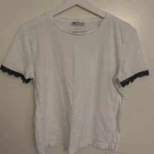 Vit t-shirt med spetsdetalj från Zara - Vit t-shirt från Zara i bomull med korta ärmar och svart spetsdetalj längst ut på ärmsluten. Enkel och stilren modell med rund halsringning, perfekt för en clean look med en twist.