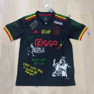 Ajax x Bob Marley Adidas fotbollströja - Svart Ajax fotbollströja från Adidas med Bob Marley-tema, färgglada ränder i rött, gult och grönt på axlar och ärmslut. Tryck med texter, illustrationer och Ziggo-logga. Material i lätt polyester, klassisk V-ringning och korta ärmar.