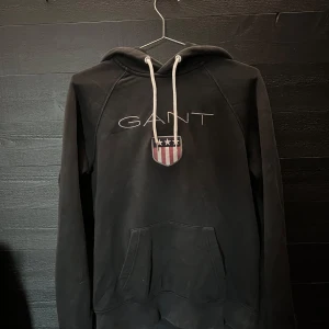 Svart GANT hoodie - Svart hoodie från GANT med broderad logga samt sköld på bröstet. Finns ett litet hål bak på ryggen.