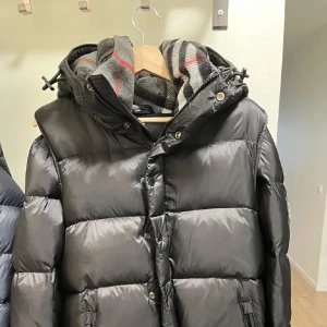 Svart dunjacka från Burberry - Snygg svart dunjacka från Burberry med klassisk puffer-design och stor huva. Jackan har dragkedja och knappar framtill samt en tydlig Burberry-logga på ärmen. Fodret är quiltat och jackan har en cool, rutig halsduk i grått, vitt och rött.
