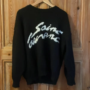 Svart stickad Saint Laurent tröja - Svart stickad tröja från Saint Laurent med vit, handskriven logga framtill. Oversized passform och långa ärmar. Perfekt för dig som gillar exklusiva och trendiga plagg med streetkänsla. Pris kan diskuteras vid snabb affär! 
