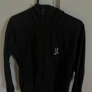 Svart vindjacka från Haglöfs med huva - Svart vindjacka från Haglöfs med hel dragkedja och justerbar huva. Jackan har diskret logga på bröstet och två fickor med dragkedja. Perfekt för blåsiga dagar och outdoor-äventyr. Snygg och funktionell design som passar till allt.