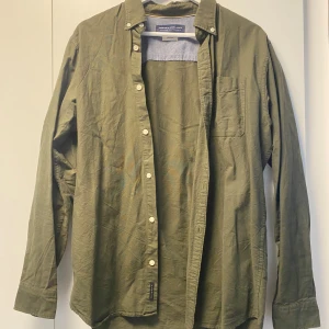 Olivgrön skjorta från Jack & Jones - Olivgrön skjorta från Jack & Jones Premium med klassisk krage och vita knappar. Skjortan har en bröstficka och långa ärmar med dubbla manschettknappar. Tillverkad i mjuk bomull som känns skön mot huden. Perfekt för dig som gillar stilren och enkel design.