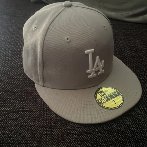 Grå LA New Era 59FIFTY keps - Snygg grå New Era 59FIFTY keps med LA-logga framtill och MLB-logga bak. Klassisk flat peak och broderade detaljer. Kepsen är tillverkad i 100% polyester och har en stilren look som passar perfekt till streetwear. Pris kan diskuteras