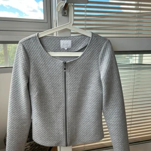 Grå cardigan jacka från Vila - Snygg ljusgrå cardigan jacka från Vila med rund halsringning och diskret strukturmönster i tyget. Jackan har lång ärm och stängs med dragkedja framtill. Perfekt för dig som gillar stilrena och enkla plagg med en twist.