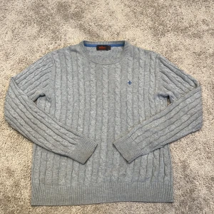 Grå kabelstickad tröja Morris M - Snygg grå kabelstickad tröja från Morris i storlek M. Tröjan är tillverkad i 100% bomull och har ett klassiskt mönster med ribbade muddar och rund hals. På bröstet finns en blå broderad logga som ger en stilren touch. Perfekt för dig som gillar clean och tidlös stil.