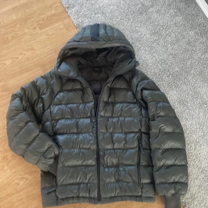 Svart dunjacka Peak Performance M - Snygg svart dunjacka från Peak Performance i storlek M. Jackan har huva, dragkedja framtill och två sidofickor med dragkedja. Klassisk pufferstil med broderad logga på bröstet och ärmen. Perfekt för kalla dagar när du vill hålla stilen.