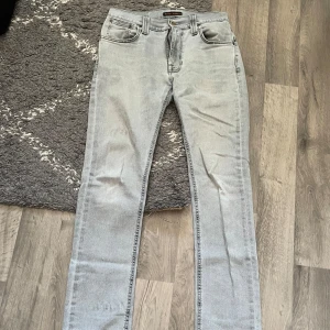 Ljusgrå jeans från Nudie Jeans - Säljer ett par ljusgrå jeans från Nudie Jeans med klassisk femficksdesign och snygga broderade detaljer på bakfickorna. Jeansen har rak passform och är tillverkade i mjukt bomullsmaterial. Perfekt för dig som gillar stilrena och tidlösa jeans.