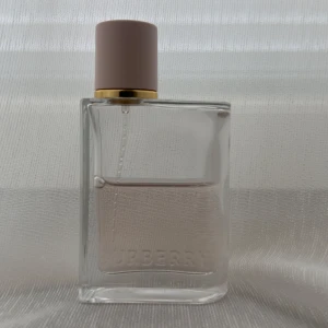 Burberry Her  - Burberry Her parfym i en stilren, rektangulär glasflaska med ljusrosa lock och gulddetalj. Flaskan är transparent med diskret rosa vätska och Burberry-loggan ingraverad framtill. Perfekt för dig som gillar eleganta och minimalistiska dofter.