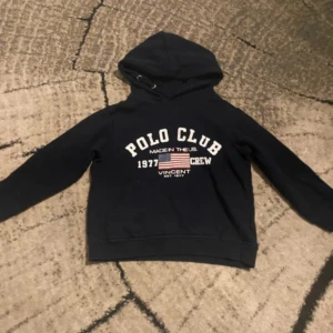 Svart hoodie Polo Club med tryck barn - Svart hoodie från Polo Club Saint Vincent med stor vit text och amerikansk flagga på bröstet. Tröjan har huva med snörning och långa ärmar. Mjuk bomullskvalitet, perfekt för chill dagar. Snygg och sportig look med crew-tryck och klassisk passform.