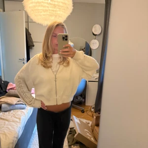 Vit croppad kofta med dragkedja - Säljer en vit croppad kofta med långärmade ärmar och snyggt hålmönster längs armarna. Koftan har en silverfärgad dragkedja framtill med rund detalj på zippen. Mjuk stickad känsla och loose passform, perfekt för lager-på-lager.