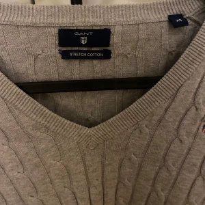 Grå kabelstickad tröja från GANT - Kabelstickad grå tröja från GANT i stretchig bomull. V-ringad modell med lång ärm och broderad logga på bröstet. Snygg ribbad kant vid hals, ärmslut och nederkant. Perfekt för dig som gillar klassisk och stilren look.